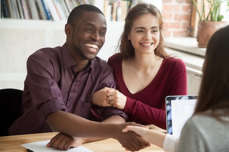 A couple smiling and shaking hands with a lady sitting behind a laptop computer